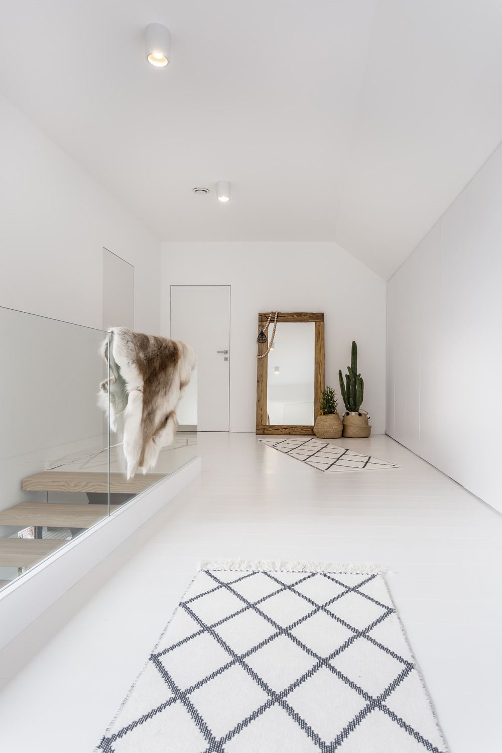 A minimalist white hallway with a geometric rug, a wooden mirror, two potted cacti, and stairs with a draped fur throw.