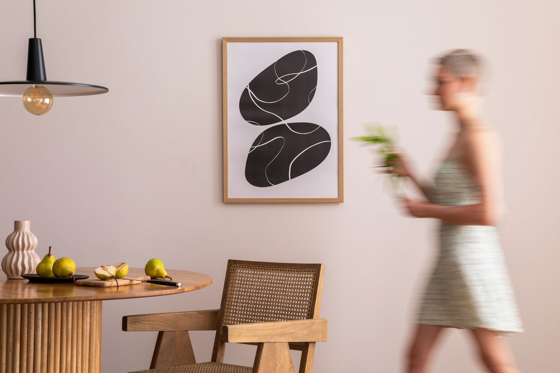 Blurred person walking past a cozy dining nook with wood table, woven chair, and framed leaf art