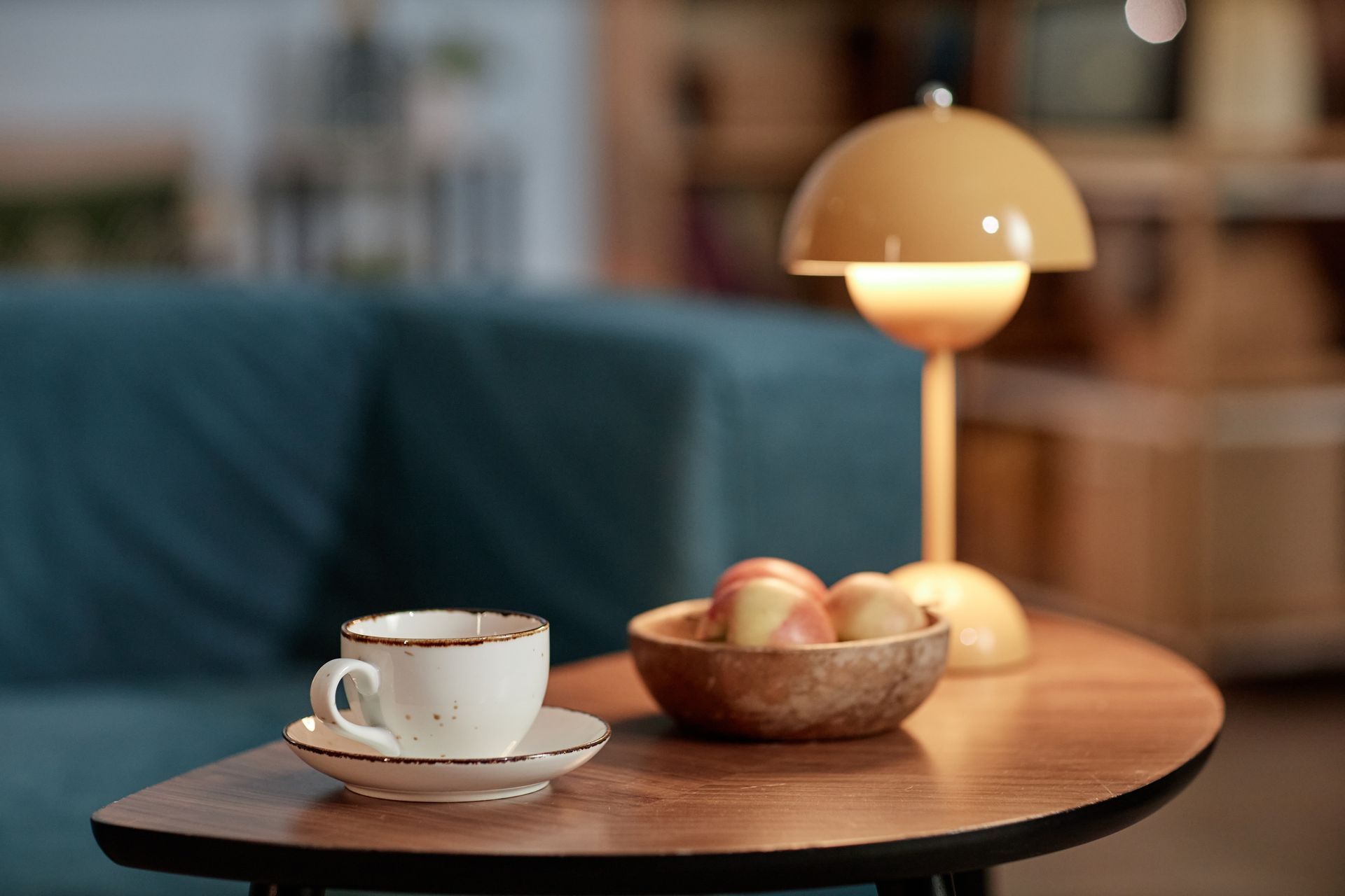 Coffee cup and bowl of apples on a wooden table beside a lit lamp in a cozy room