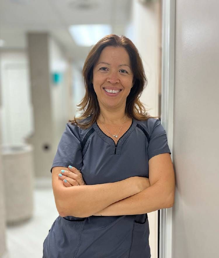 A woman in a scrub is leaning against a wall with her arms crossed and smiling.
