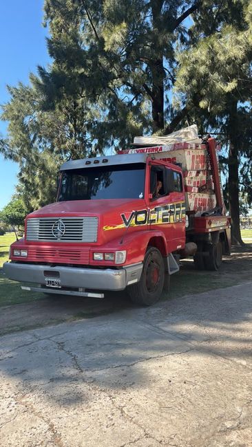 Camión volquete rojo aparcado en un camino de tierra bajo los árboles a plena luz del día.