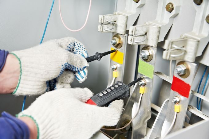 Un operaio con i guanti da lavoro utilizza un tester di tensione per controllare la corrente sulle sbarre collettrici elettriche contrassegnate da etichette gialle, verdi e rosse.