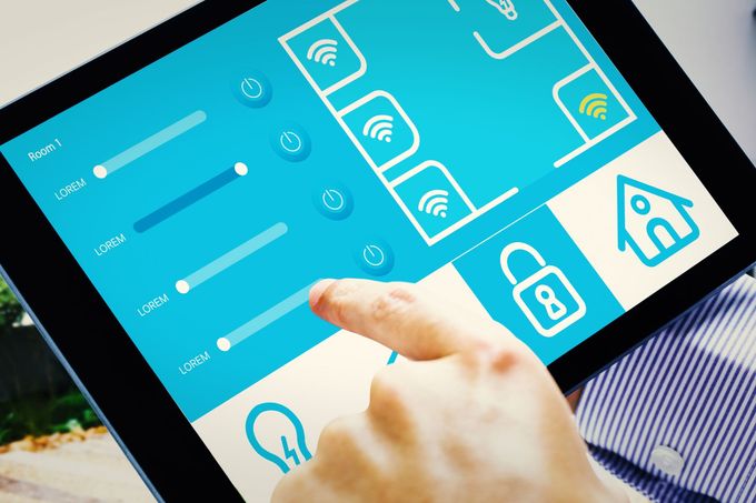 Una mano tocca lo schermo di un tablet che mostra un'interfaccia di controllo per la casa intelligente con cursori, pulsanti di accensione e icone delle stanze.