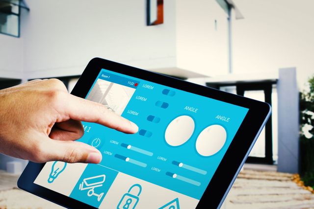 Una mano tocca lo schermo di un tablet che mostra un'interfaccia blu per il controllo della domotica, davanti a una casa moderna.