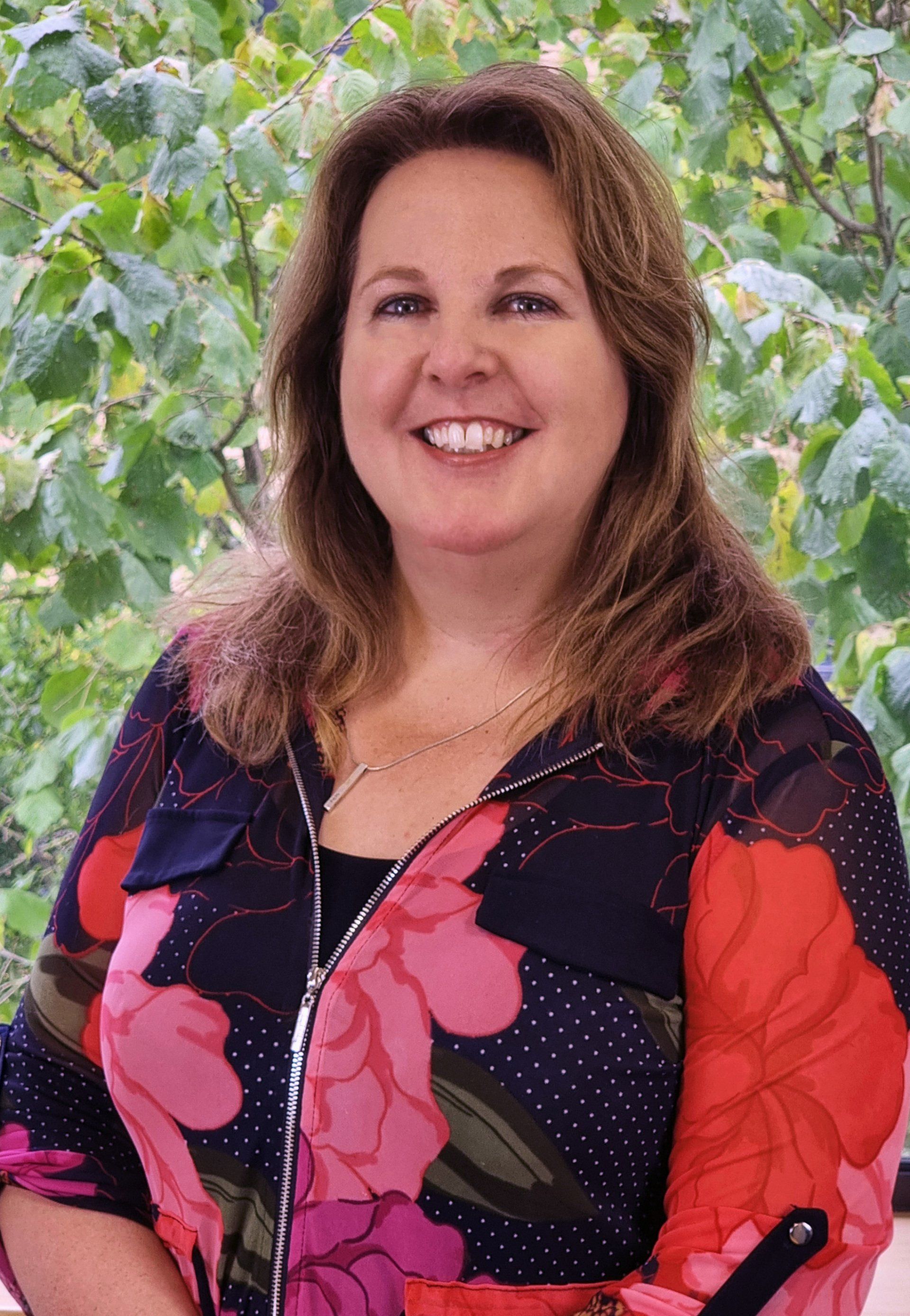 A woman in a floral shirt is smiling in front of a tree.