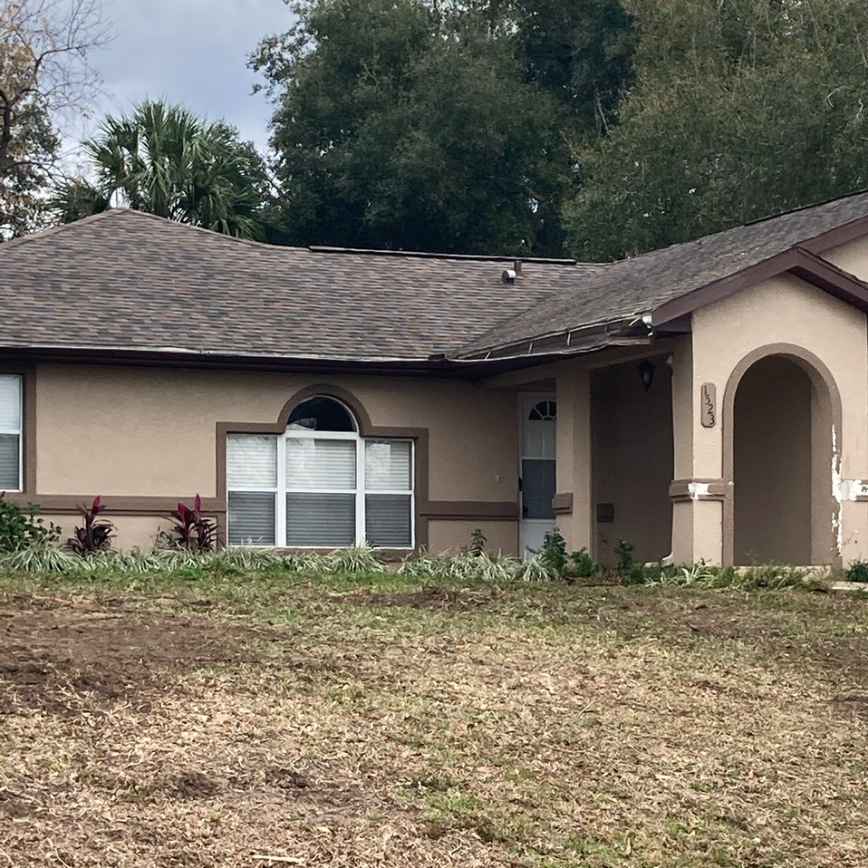 Single-story beige house with a brown roof and arched front entry, seen from a dry front lawn.