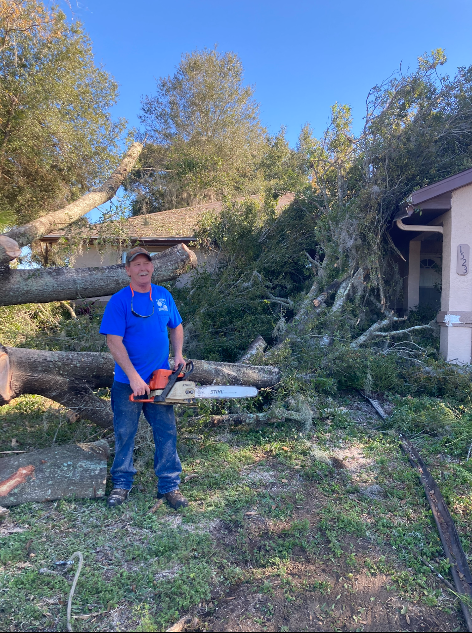 Man in blue shirt holding a chainsaw beside a fallen tree in a yard after storm damage