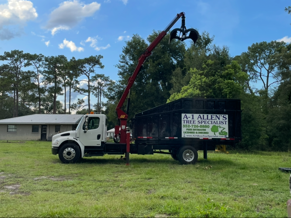 White truck with red crane arm loading debris into black dump truck in a grassy yard.