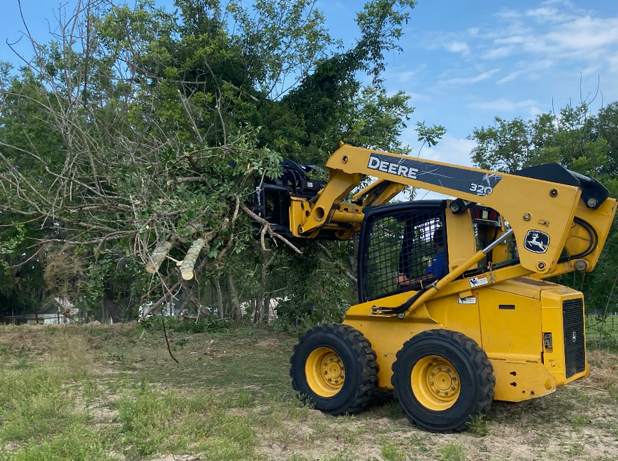 Yellow John Deere skid steer lifting tree branches in a grassy field under a blue sky