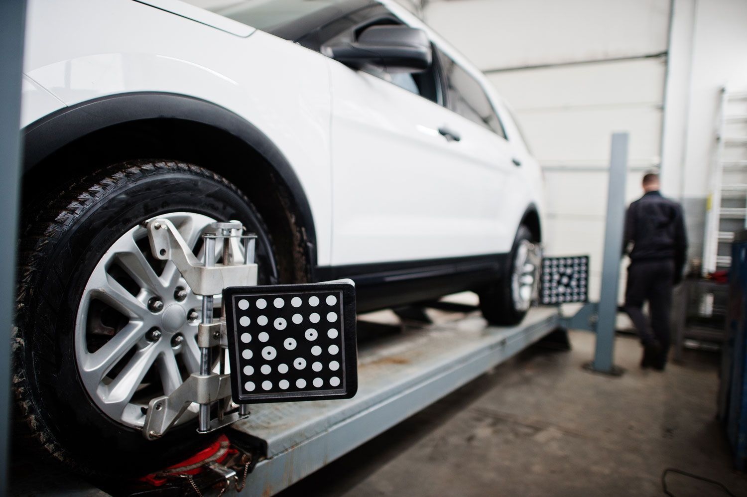 White SUV on wheel alignment rack in auto shop, with technician in background