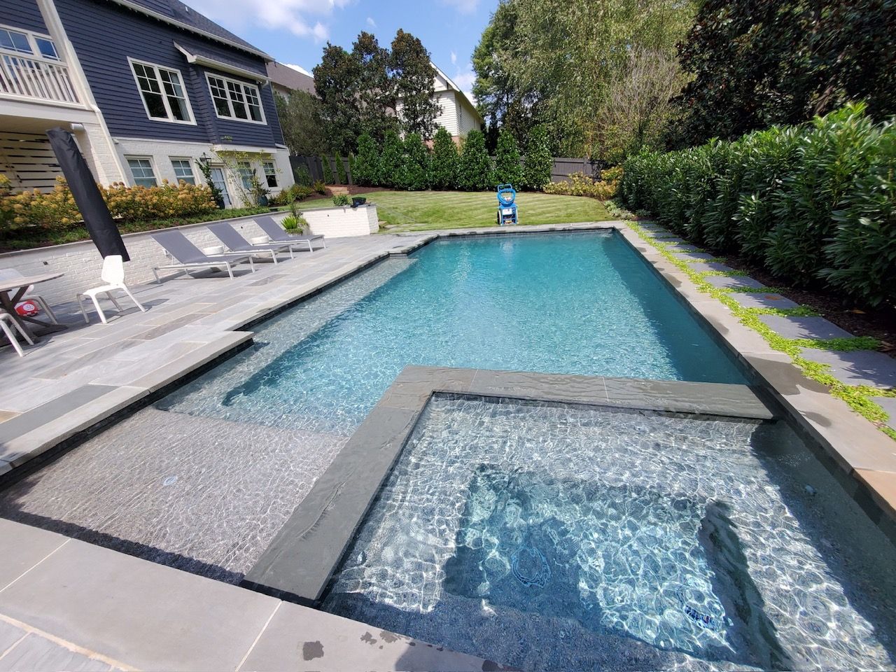 Backyard swimming pool with hot tub, lounge chairs, and modern house beside lush greenery