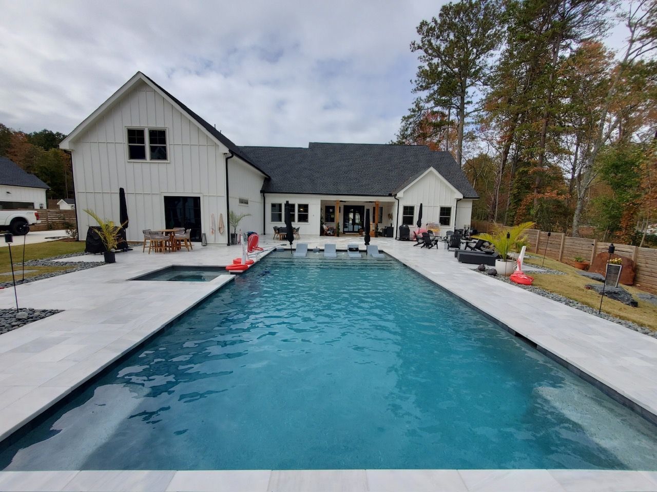 Backyard swimming pool beside a modern white house with patio chairs and trees in fall.