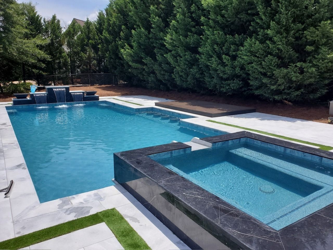 Backyard pool with attached hot tub, blue water, and white deck bordered by tall green hedges
