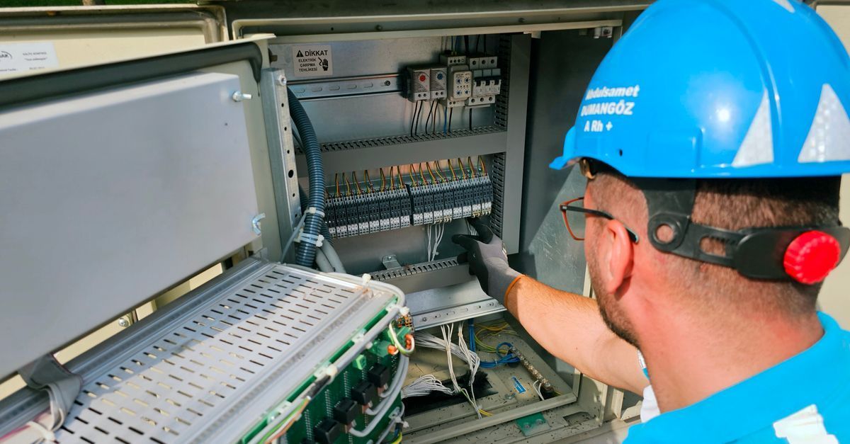 Técnico con casco azul inspeccionando un panel de control eléctrico con las puertas del armario abiertas