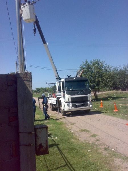 Camioneta de servicios blanca y trabajadores reparando líneas eléctricas junto a la carretera, cerca de conos naranjas, en una vía rural.