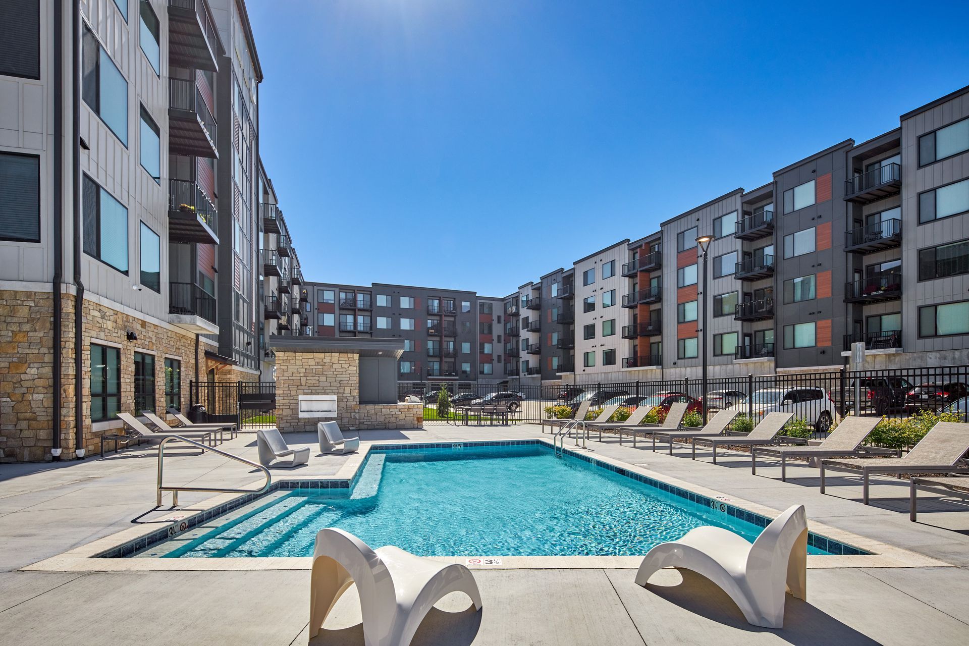 Pool area at a modern apartment complex, featuring a pool, lounge chairs, and buildings under a sunny sky.
