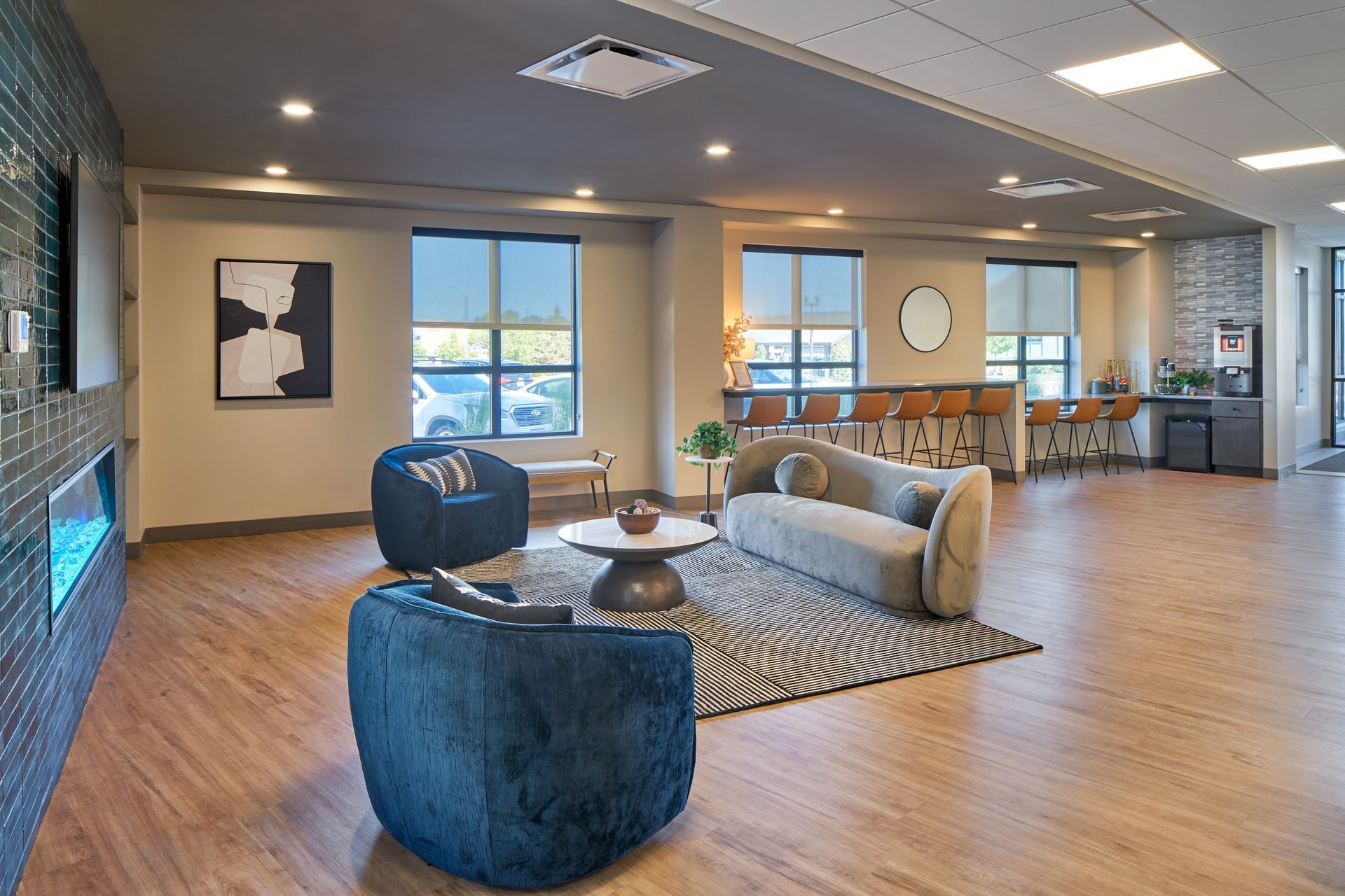 Spacious modern lounge with seating, coffee table, and bar area. Light wood floors, windows, and decorative art.