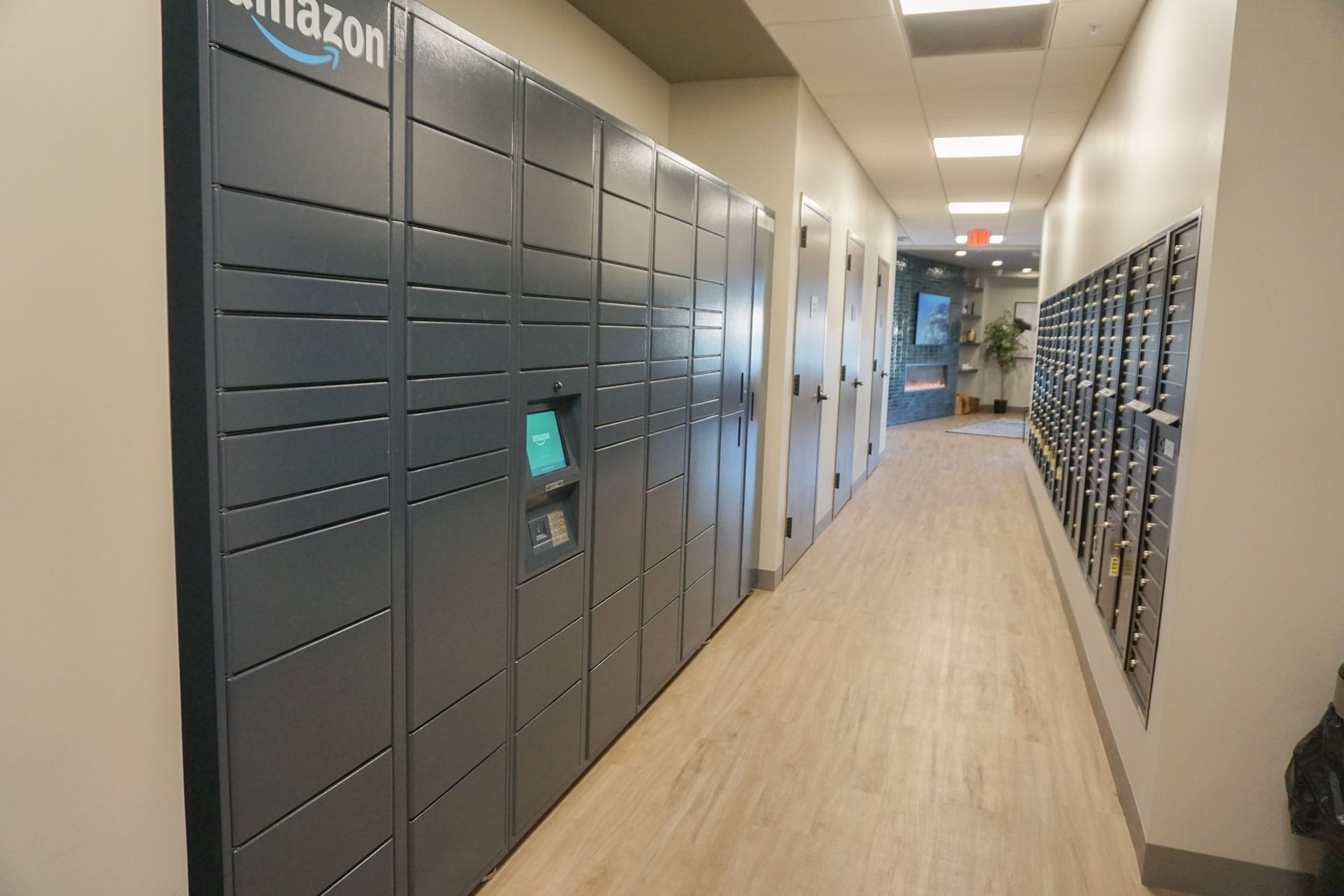 Amazon lockers and mailboxes in a hallway. The lockers are dark gray, and the hallway has wood flooring.