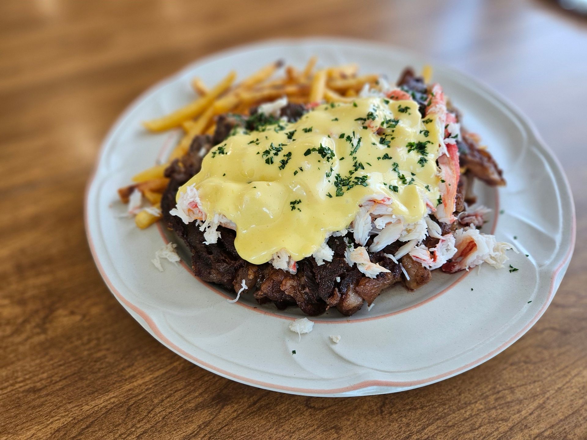 Plated burger topped with hollandaise and herbs, served with fries on a white plate at a wooden table