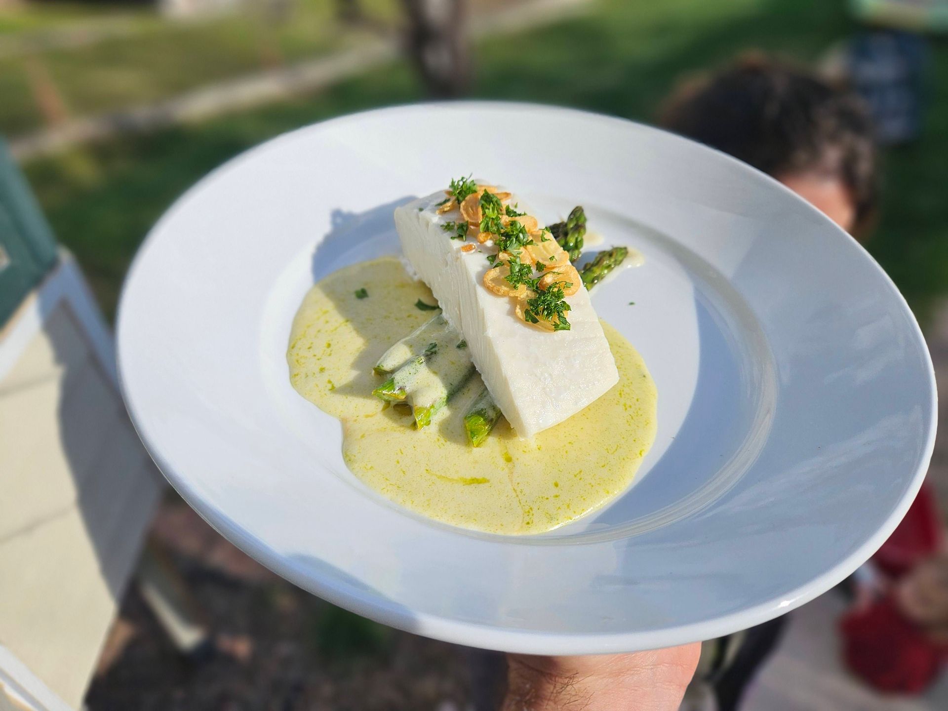 White plated gourmet dessert with a creamy sauce and herb garnish outdoors, held in hand