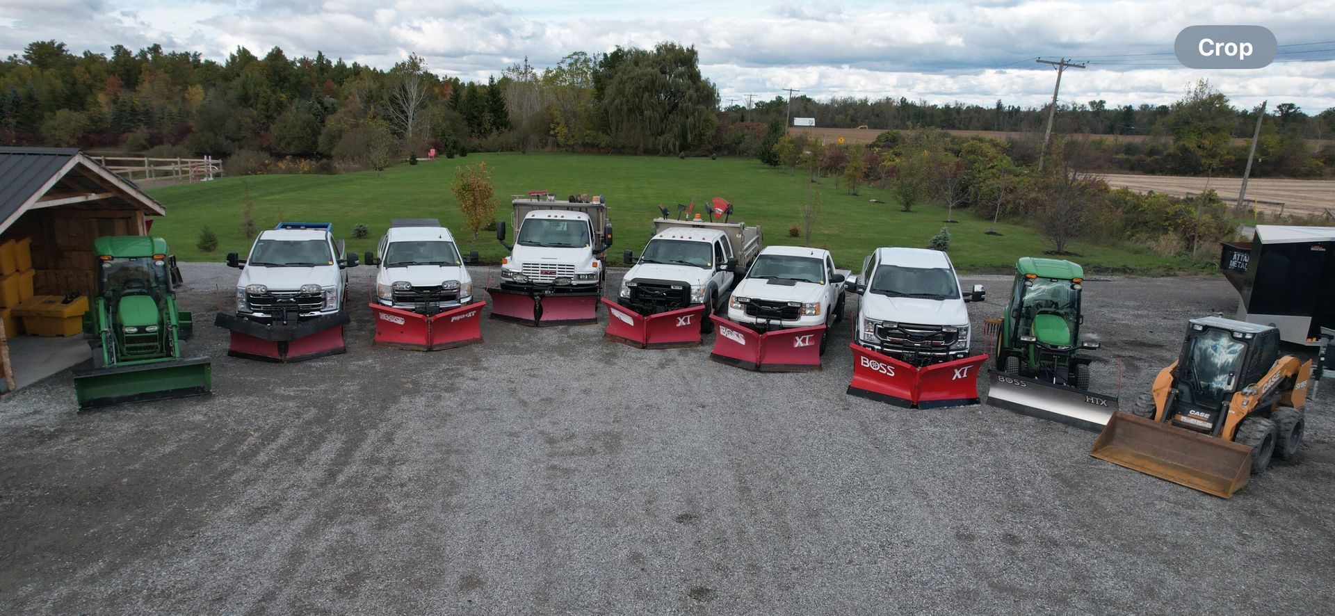Vehicles with snow plows lined up on gravel, in front of a field and trees, under an overcast sky.