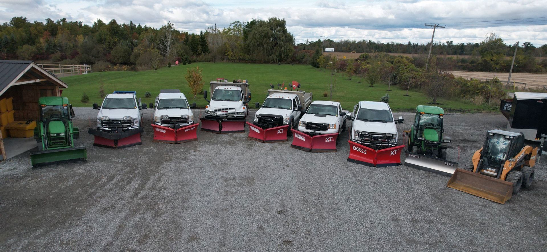 Snow plows lined up on a gravel lot; green tractors, white trucks with red plows, and a skid steer.