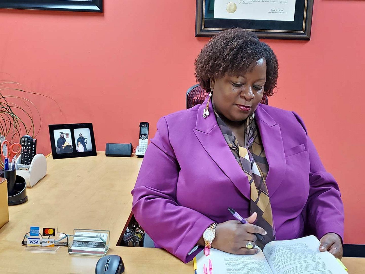A woman in a purple jacket is sitting at a desk reading a book.