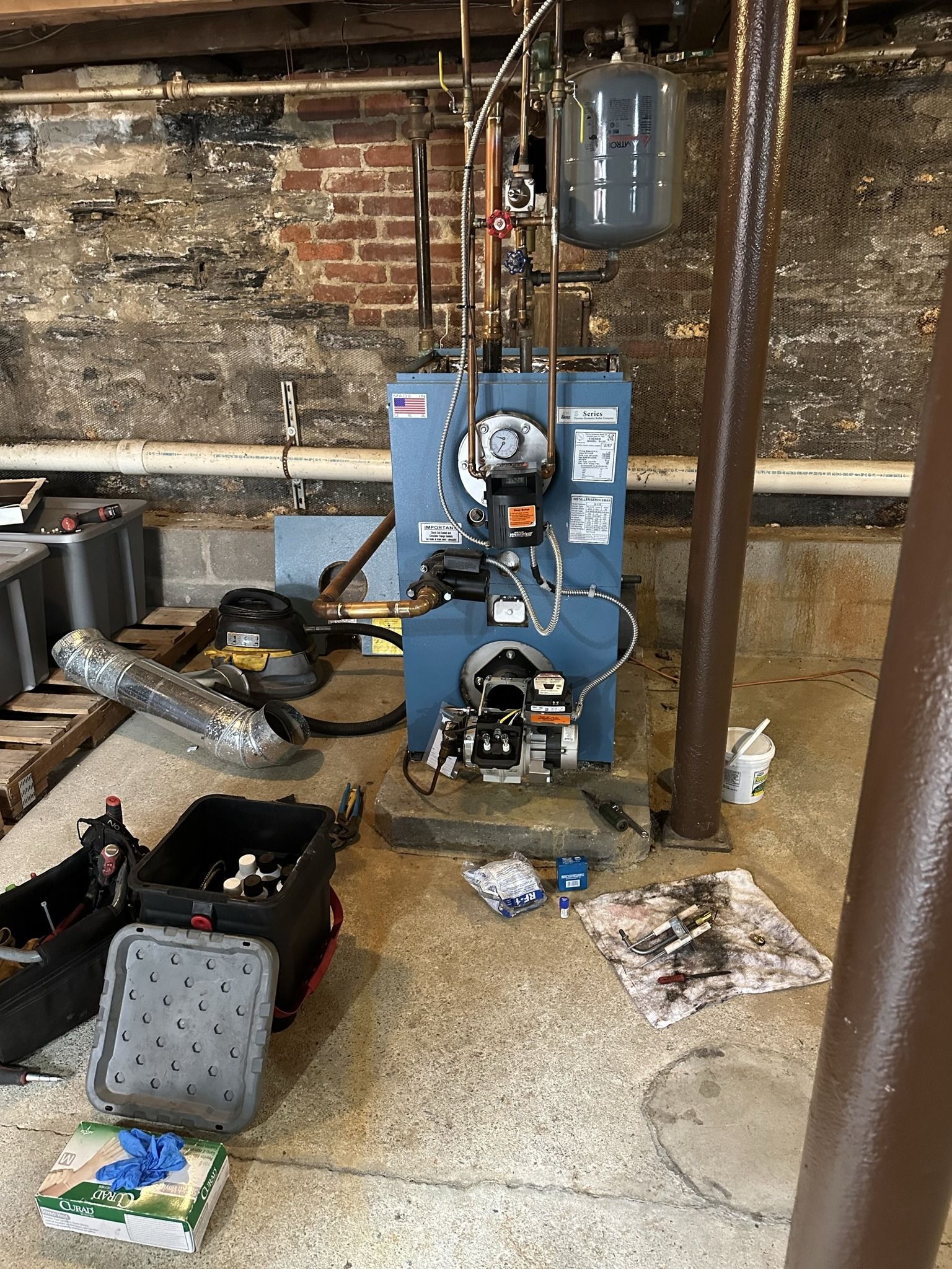 A blue oil-fired boiler stands on a concrete floor in a basement, surrounded by various tools, parts, and maintenance gear.