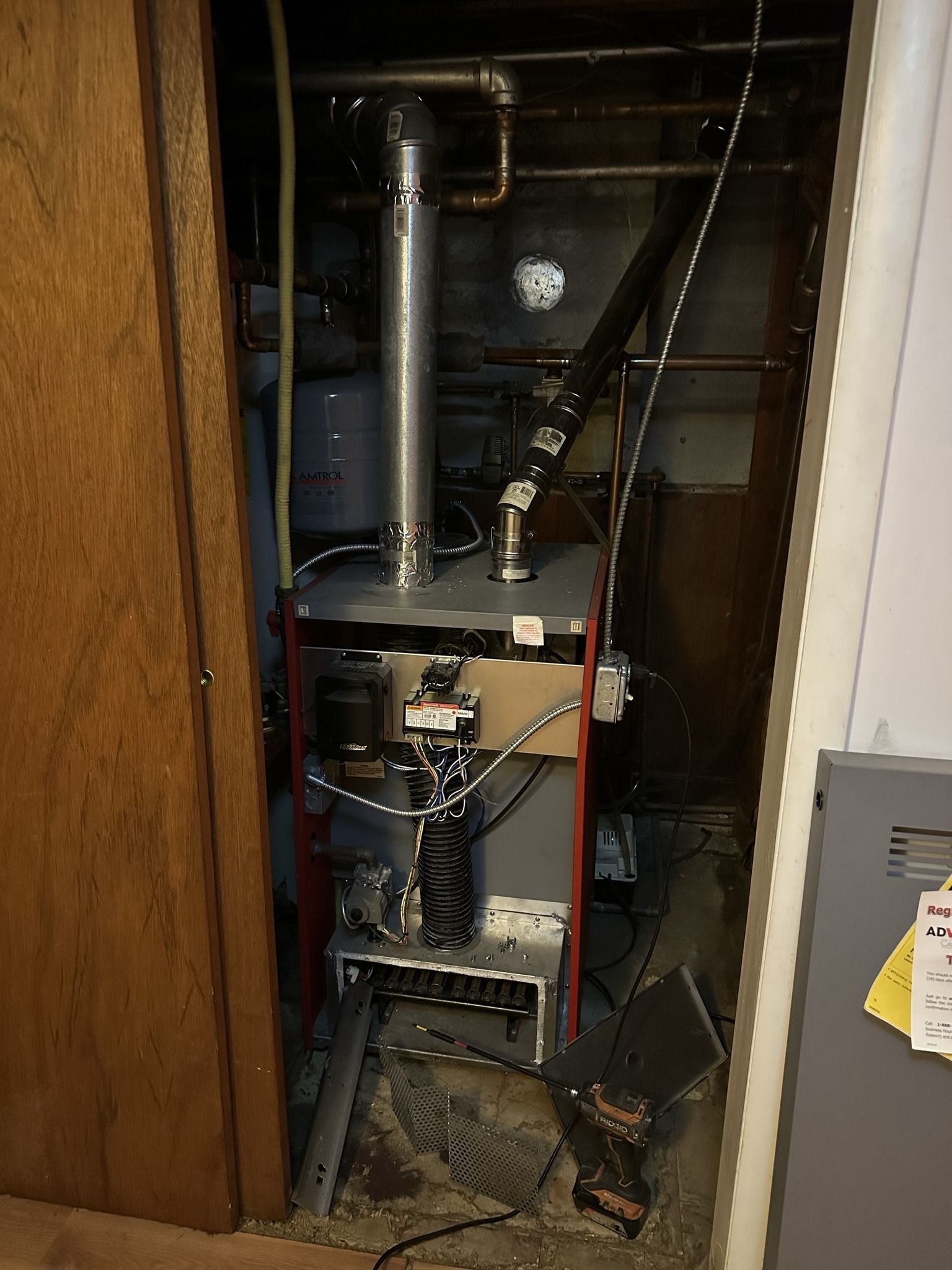 A red-trimmed oil furnace situated in a dimly lit utility closet, with metal venting and plumbing visible above it.