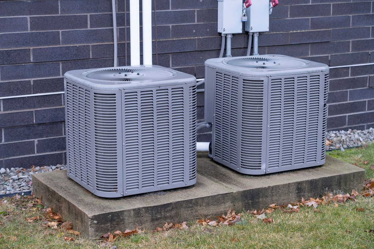 Two grey residential air conditioning units sit on a concrete pad against a dark brick wall.