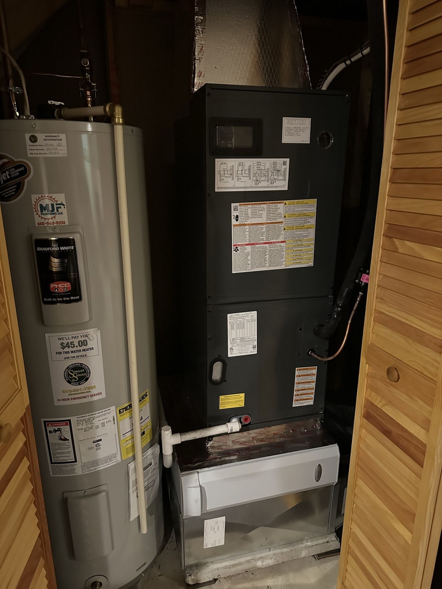 A water heater and a furnace unit standing side by side in a narrow, wood-paneled utility closet.