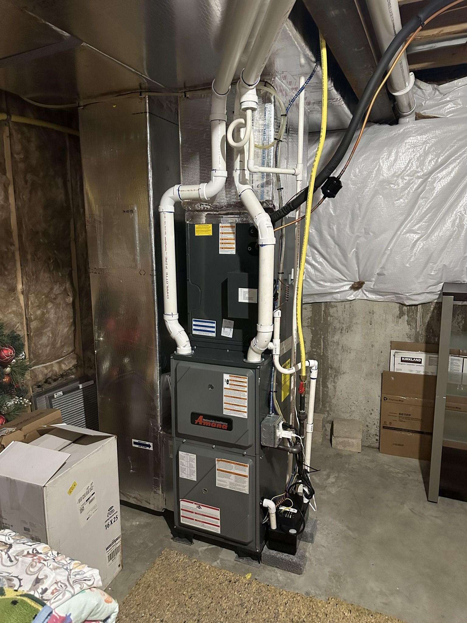 A tall, gray, multi-section furnace unit stands in a basement with attached white PVC venting pipes and metallic ductwork.