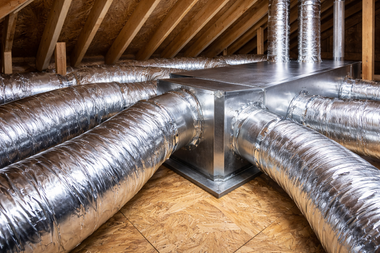 A central metal HVAC distribution box with multiple silver insulated flexible ducts in an unfinished attic space.