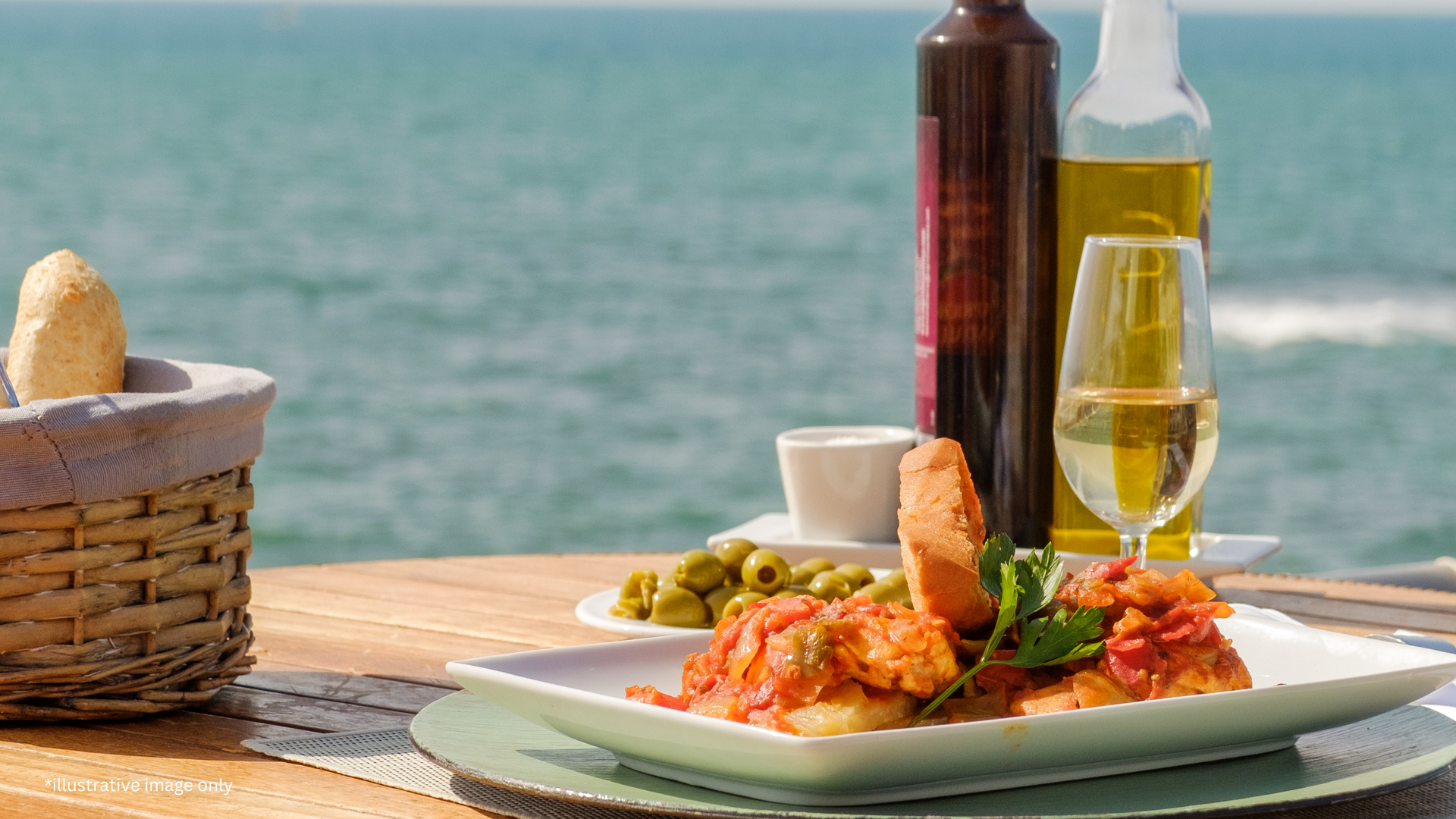 A plate of food and a glass of wine on a table with the ocean in the background.