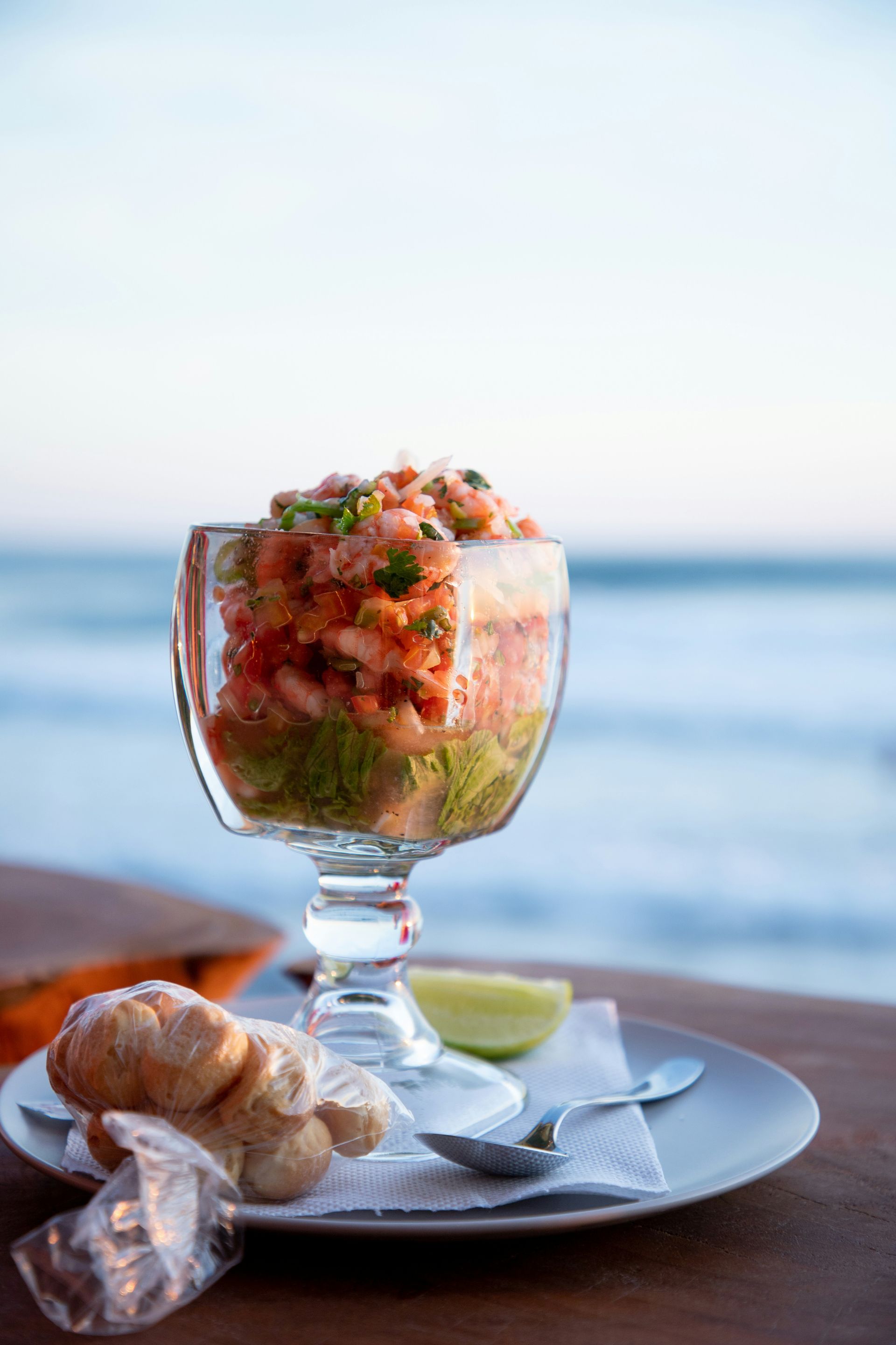 A glass filled with shrimp and vegetables is on a plate next to a spoon.