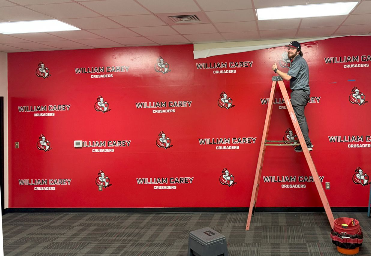 A person stands on a tall ladder, applying red wall decals with logos to a large, bright red wall in an office space.
