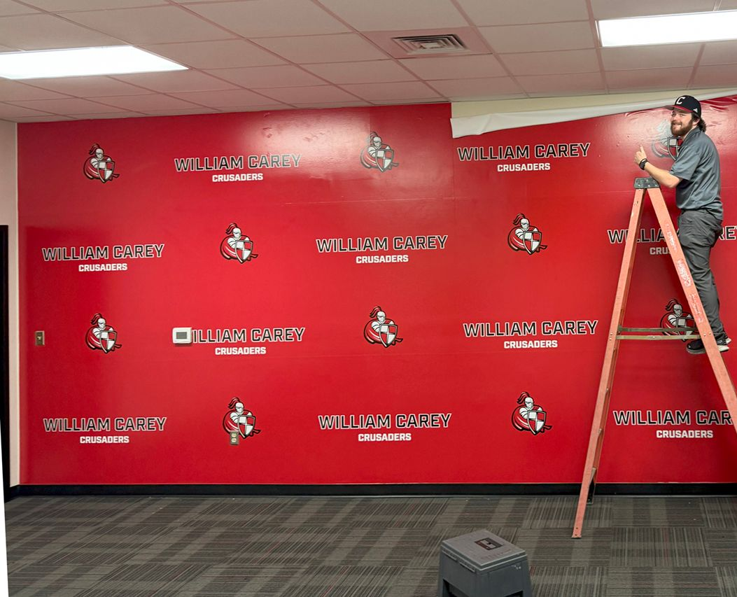 A person on a ladder installs a red wallpaper featuring a repeating pattern of logos and text on an office wall.