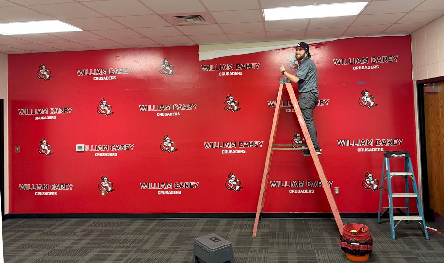 A person on a ladder installs a red adhesive wall mural featuring a repeating logo and text in an office setting.