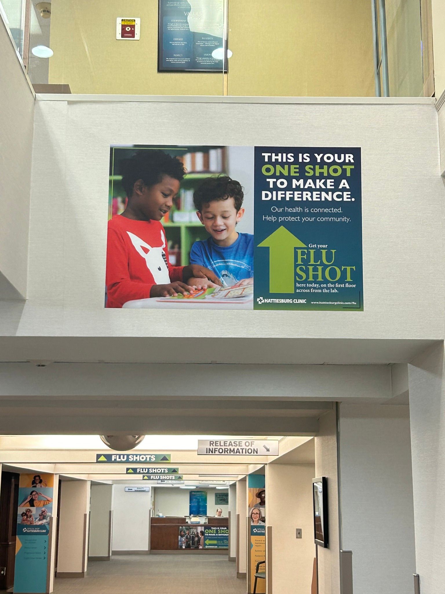 A hallway sign advertising flu shots with an image of two people at a table, featuring a large green arrow pointing upward.