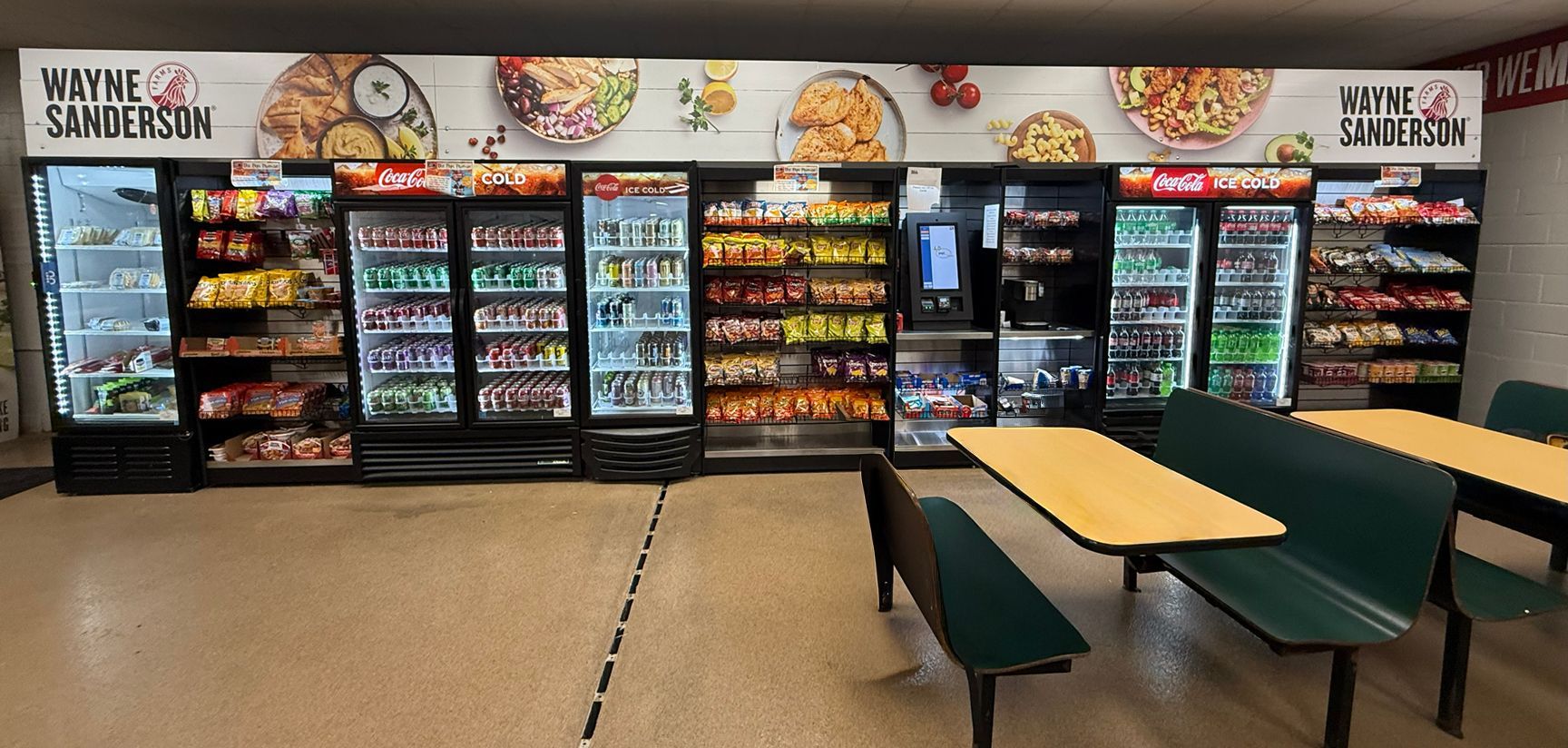 A breakroom with rows of vending machines stocked with drinks and snacks, alongside tables and benches for seating.