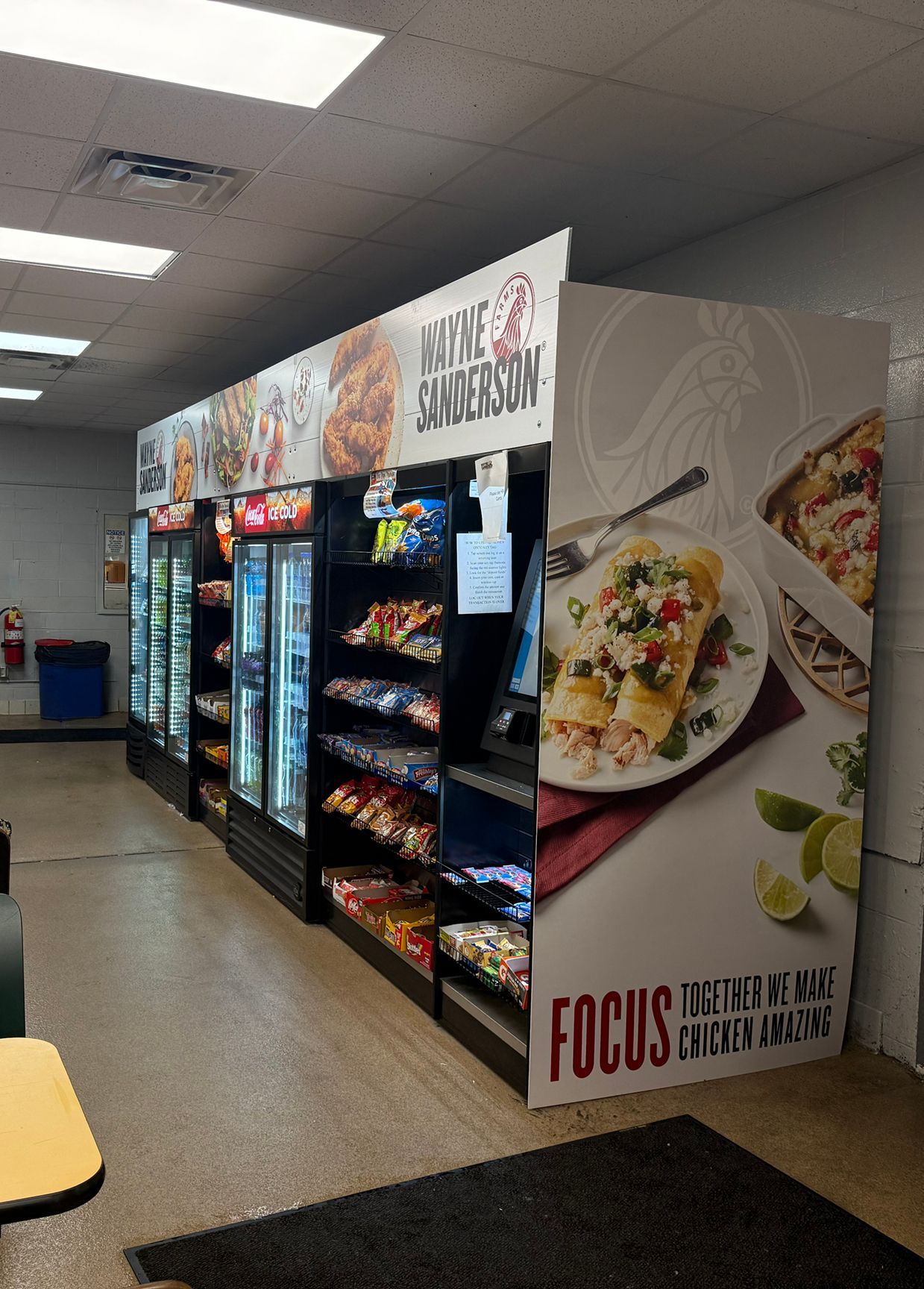 Retail display with glass-door coolers and shelves of snacks, topped with food branding posters in a brightly lit room.
