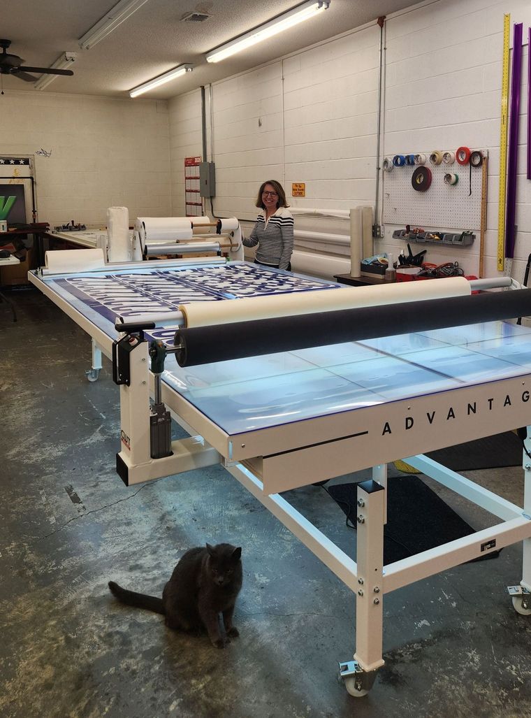 A person stands behind a large quilting machine in a workshop, with a dark cat sitting on the concrete floor in front.