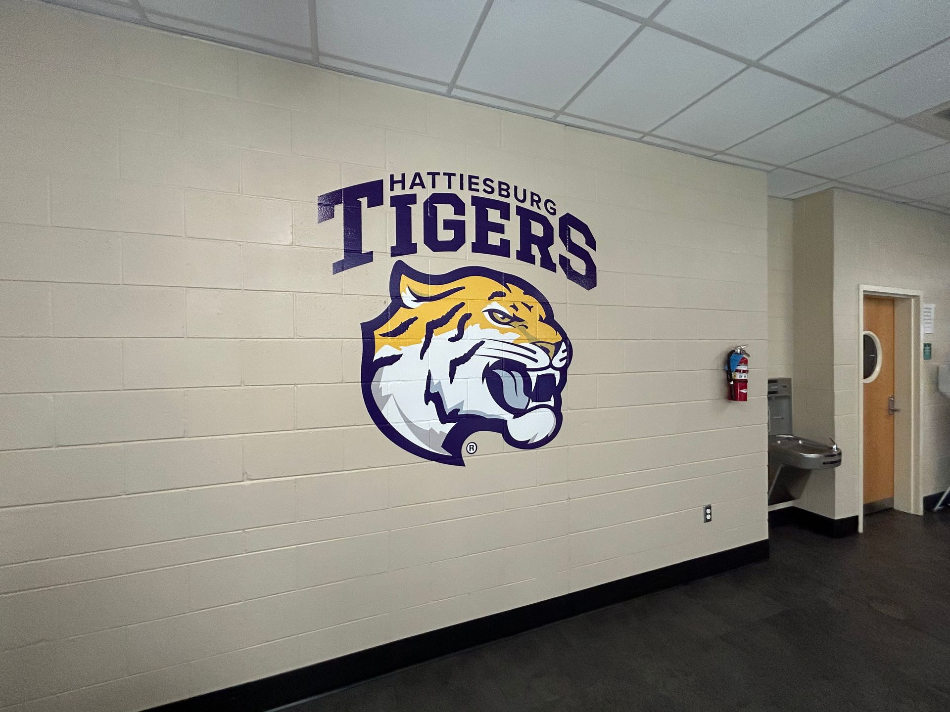 A purple and gold Hattiesburg Tigers logo painted on a cream-colored concrete block wall inside a school building.