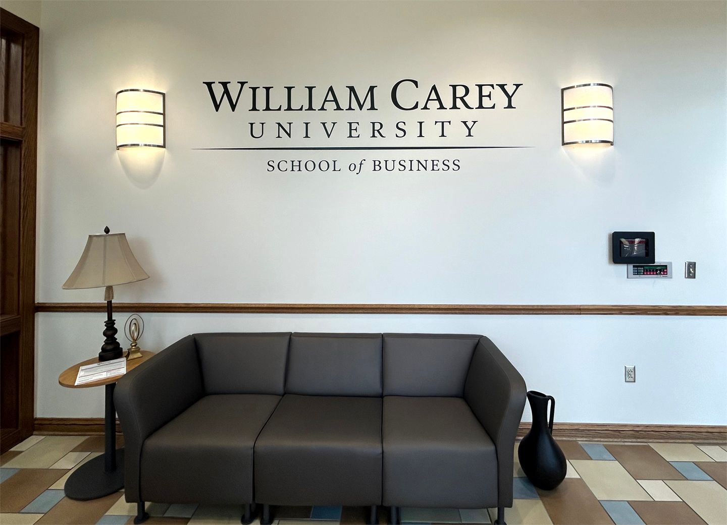A waiting area with a gray couch, side table with a lamp, and wall signage for the William Carey School of Business.