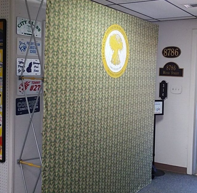 An office wall with a patterned green tapestry, a gold church logo, a metal sign display, and room number plaques.