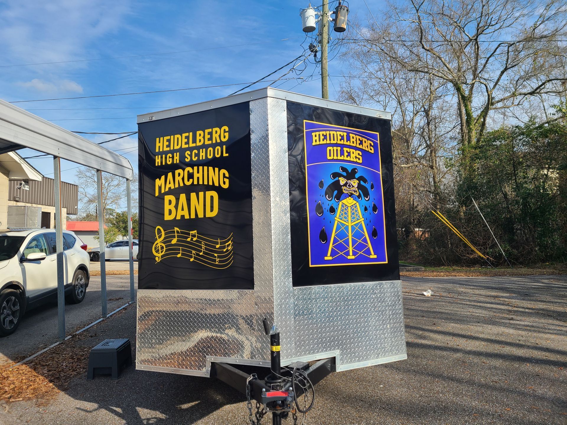 A silver cargo trailer features signage for the Heidelberg High School Marching Band, parked outdoors in a gravel lot.