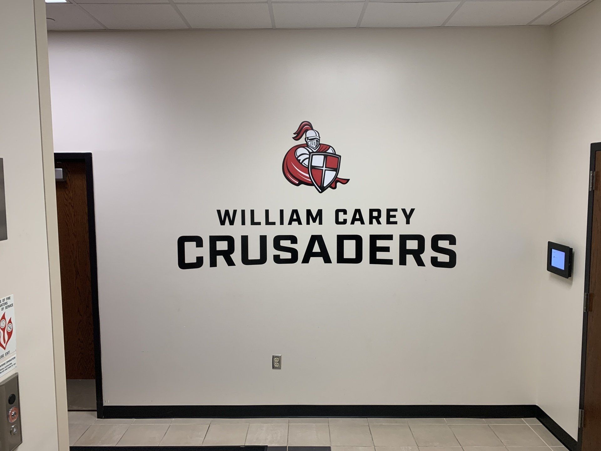 Wall decal with the William Carey Crusaders logo and text in black and red on a white interior wall.