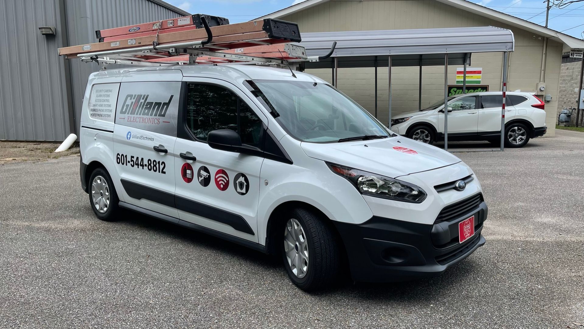 A white Ford Transit Connect service van with ladder racks parked on a gravel lot in front of a metal building.