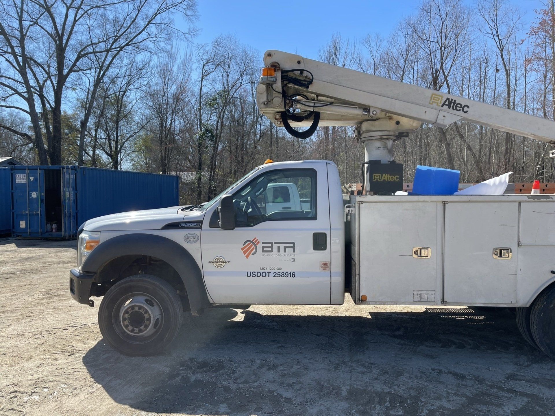 A white utility truck with an Altec crane lift parked on a gravel lot next to a blue shipping container.