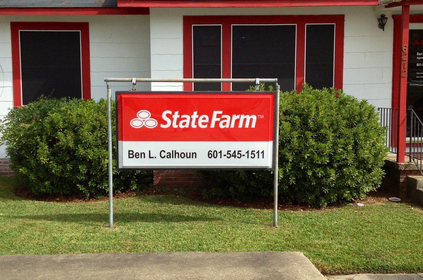 A State Farm sign for agent Ben L. Calhoun stands in front of a white building with red trim and two green bushes.